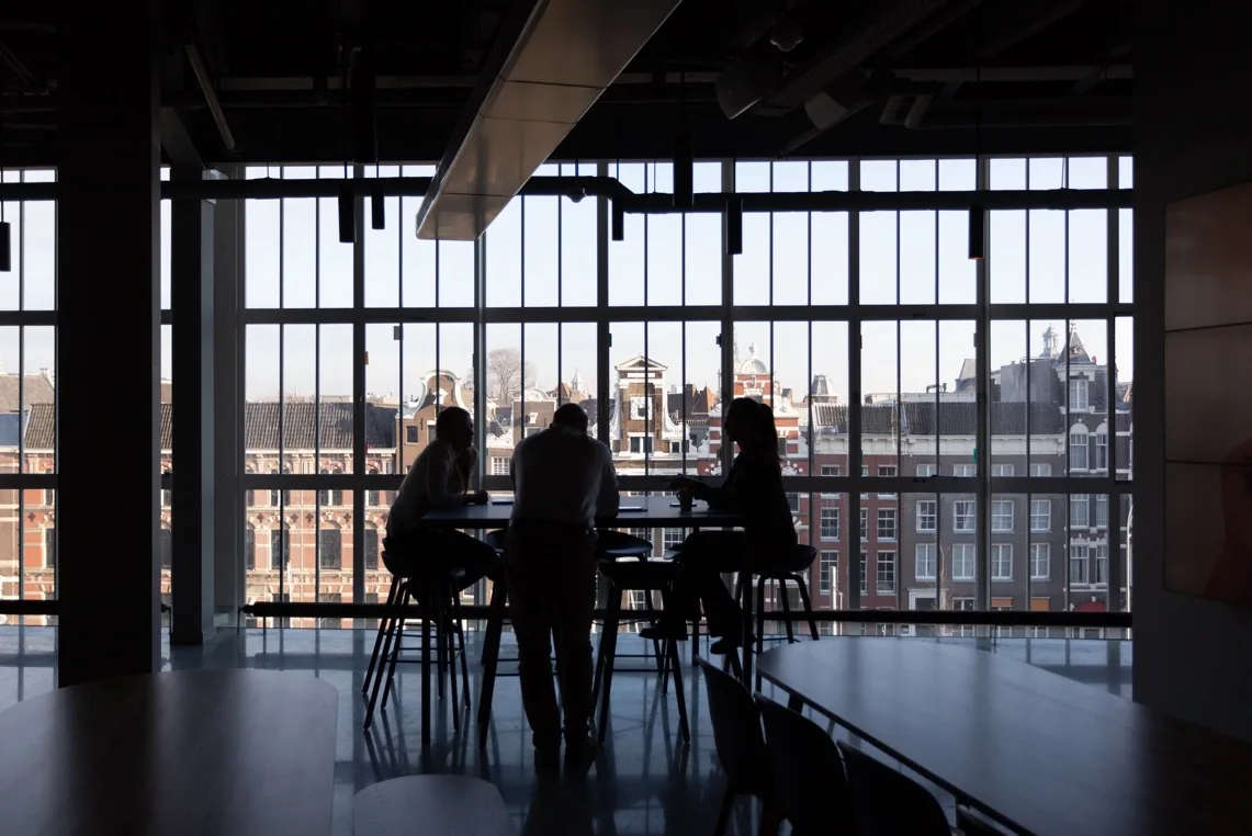 Silhouetted figures at a table backlit by tall windows overlooking a historic city skyline.